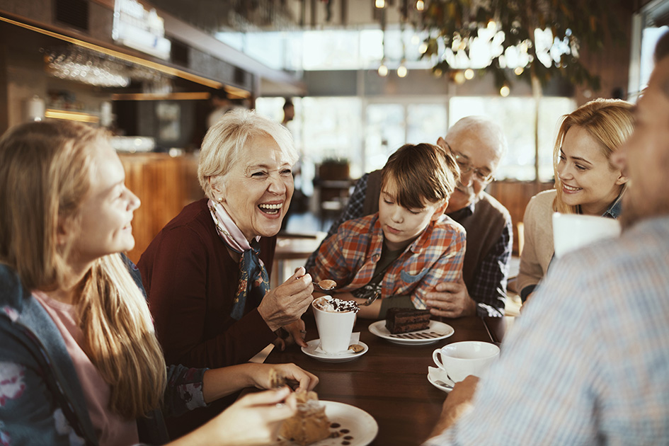 Eine große Familie stitzt in einem Restaurant und unterhält sich ausgelassen. Die Oma in der Mitte lacht fröhlich in die Runde.