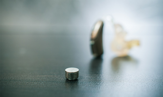 Hörgerätbatterie liegt auf einer scharzen Fläche. Im Hintergrund erkennt man unscharf ein Hörgerät stehend