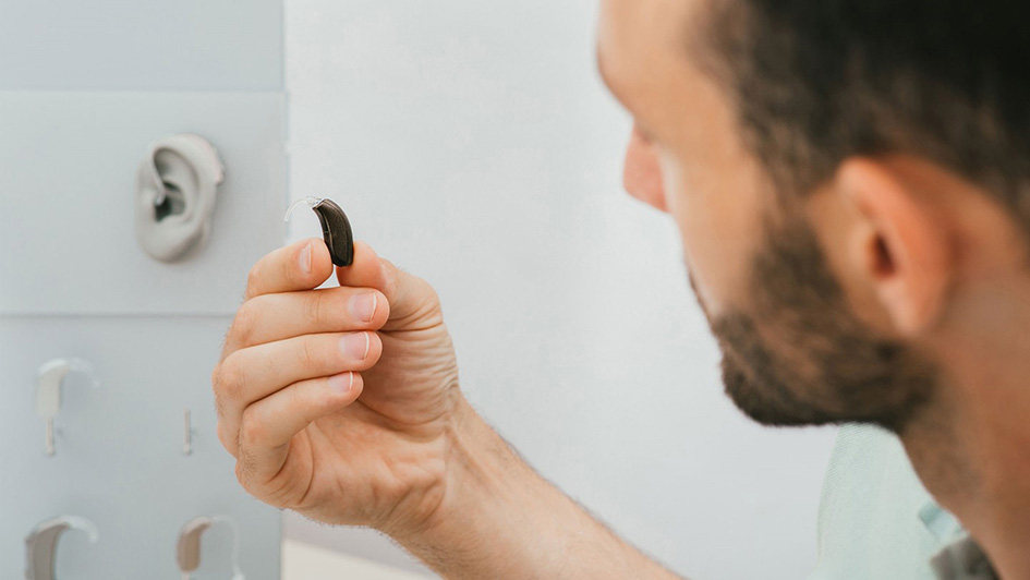 Mann hält ein Hörgerät in der Hand, im Hintergrund sieht man ein verschiedenen Modell