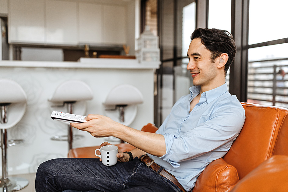 Junger Mann mit Hörgerät sitzt mit Kaffe auf der Couch und schaltet durchs TV-Programm